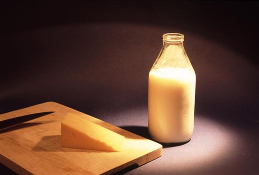 Glass bottle full of fresh creamy cows milk and a wedge of cheddar cheese on a cheeseboard over a dark background with copyspace