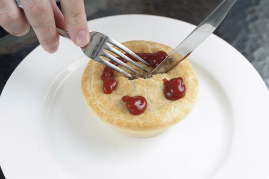 Man eating a freshly baked golden meat pie for lunch with a smiling gravy face on the pastry crust, high angle view as he cuts into it with a knife on a white plate