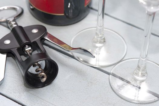 Lever style corkscrew bottle opener alongside wineglasses and a bottle of wine on a white wooden table