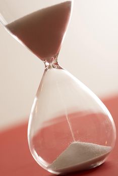 Close up cropped angled view on large glass hourglass with sand pouring close up over red surface