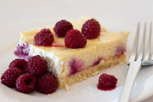 Slice of baked ricotta cheesecake topped with fresh ripe raspberries with a serving of raspberry compote on the side