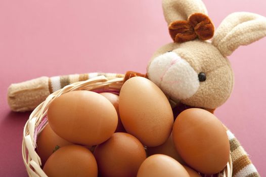 Cropped close up on bunch of brown eggs in basket with bunny doll over muted pink background
