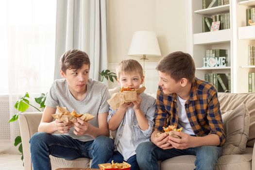 Three cute teenagers, in casual clothes, are sitting on the sofa in the living room, eating fast food, a hamburger, communication