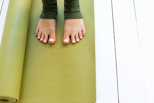 Female beautiful feet in green leggings on green rubber sports mat or fitness mat lying on white wooden floor. Copy space