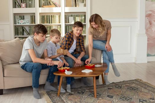 Happy family, a woman and three teenage boys, sit on the sofa in the living room, eat fast food, in red boxes. Copy space