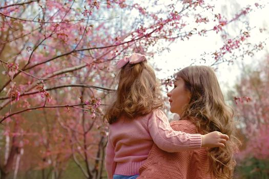 Mother and little daughter, standing in the park under a flowering cherry tree. Back view. copy space