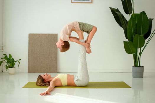 A beautiful slender woman, doing yoga with a child boy, lies on a sports mat, raising her legs up, doing exercises with support. copy space.