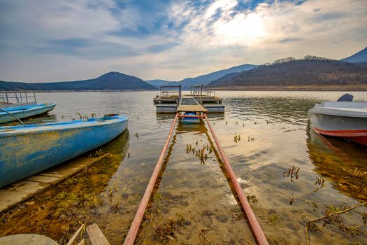 The tranquil afternoon on a lake with a wooden pier and boats in the colorful sky.