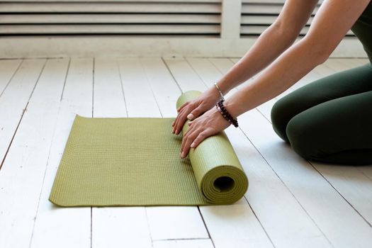 Young slender woman's hands twisting green mat for fitness or yoga on white wooden floor. Close up