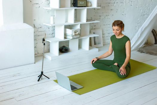 A beautiful and slim woman in green sportswear writes down yoga classes at home, in a light interior, in front of a laptop and a smartphone on a tripod