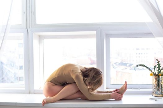 A slender European woman in a beige swimsuit performs a yoga exercise, near a large white window. copy space