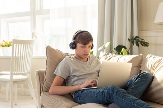 A teenager boy in a gray t-shirt and blue jeans, lying on a sofa in a room, wearing black headphones on his head, looks into a gray laptop, on his knees. Copy space