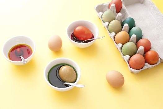 Process of dyeing Easter eggs in white bowls with liquid paint, spoons and box of colored eggs on yellow table surface background