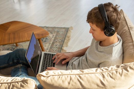 A teenager in a gray T-shirt and blue jeans, sits on a sofa in a room, wearing black headphones on his head, plays a computer game on a laptop. Top view