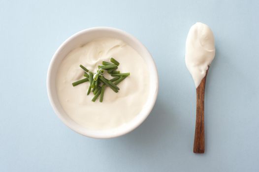 Bowl of sour cream and chopped fresh chives with a used dirty wooden spoon alongside, overhead view on blue