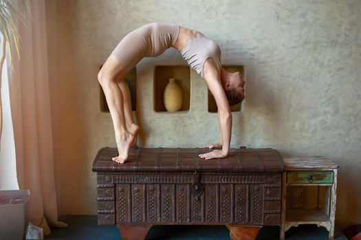 A beautiful and slender woman in a beige top and breeches, stands in a yoga pose, performs the exercise -bridge, in a room with an oriental interior