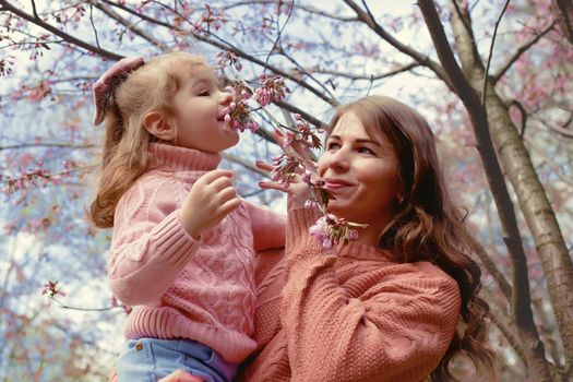 Mother and little daughter, standing in the park under a flowering cherry tree, inhale the aroma of flowers