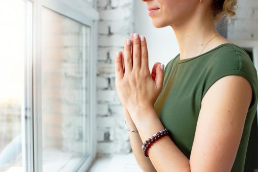 Girls in a green T-shirt, in a yoga pose close-up, folded their palms together on their chest, close-up, stands by the window in the daylight