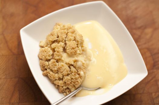 Tasty apple crumble dessert served with custard in a modern square pudding bowl on a wooden table