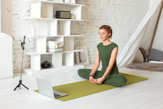 A slender beautiful woman in a green sports jumpsuit, in a bright room, is broadcasting online, performs a yoga exercise, there is a laptop and a smartphone on a tripod nearby