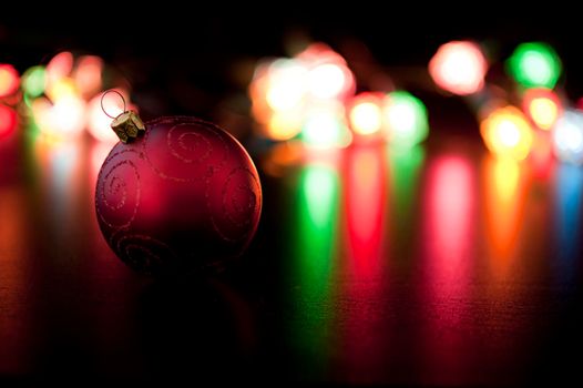A single red bauble decoration lies in front of a background of colourful Christmas lights shining across a reflective surface