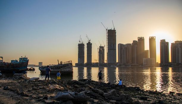 Sunrise in Jadaf area of Dubai, view of Dubai creek Harbor construction of which is partially completed. Old abandoned ships can be seen on the scene. Some men fishing. Outdoors