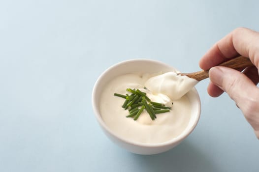 Man dipping a small spoon into a white ceramic bowl of sour cream garnished with fresh chopped chives over a grey background with copy space