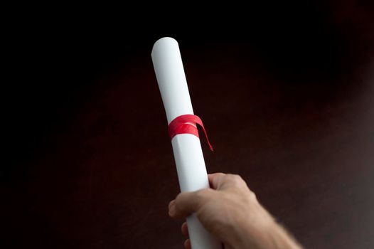 Studio shot over black of a a male hand holding a diploma or document, rolled and tied with red ribbon