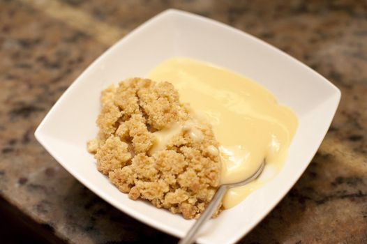Serving of delicious apple crumble and custard for dessert in a modern square white bowl, high angle view of the crumbly topping