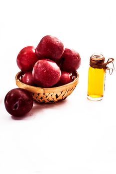 Raw organic red ripe plums in a brown-colored basket along with its extracted essential oil in a transparent glass bottle isolated on white.