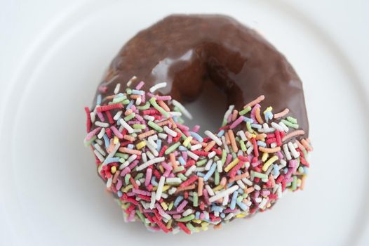 Freshly baked single chocolate doughnut dipped in multicolored candy sprinkles served on a white plate for a morning coffee break