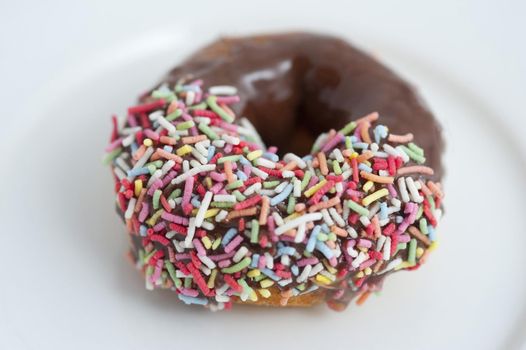 Chocolate glazed ring donut dipped in colorful candy sprinkles on one side on a white plate with focus to the sprinkles