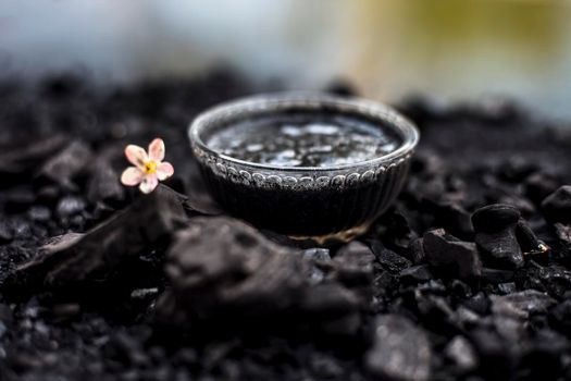 Close up of activated charcoal in a glass bowl on the wooden surface along with some raw powder of charcoal or coal spread on the surface.