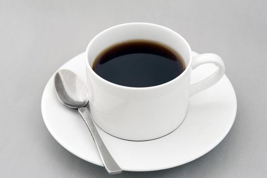 Cup of strong black espresso coffee served in a generic white ceramic cup and saucer with teaspoon over a grey background