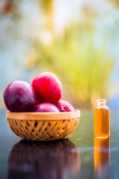 Raw organic red ripe plums in a brown-colored basket on the wooden surface along with its extracted essential oil in a small transparent bottle with blurred background.