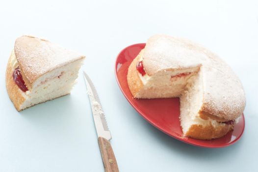 Freshly baked jam sponge cream cake with a generous slice on the side alongside a knife, served on a red plate