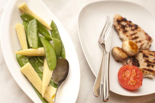 Serving of two lean grilled pork chops with mushrooms and tomato and a side dish of vegetables containing mangetout or sugar snap peas and baby corn on the cob
