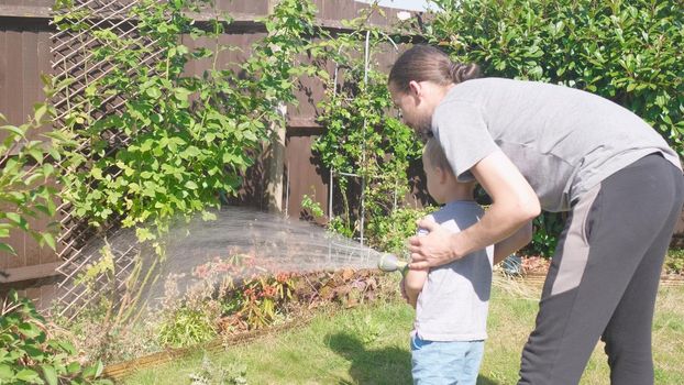 Father and Son. Funny little boy watering lawn plants in garden housing backyard with Dad. Summer house work. Hardworking preschool Kid outdoors. Children help with housework. activity for kids.