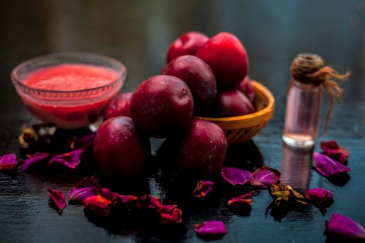 Raw organic red ripe plums in a brown-colored basket along with its extracted essential oil in a transparent glass bottle along with its paste in another glass bowl.