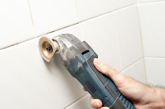 Man removing grout or old cement between white wall tiles with an oscillator in a DIY concept