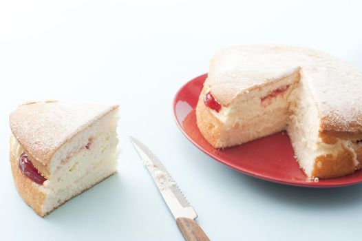 Fresh Victoria sponge cake with strawberry jam and cream served on a red plate with a single large slice cut and placed to the side