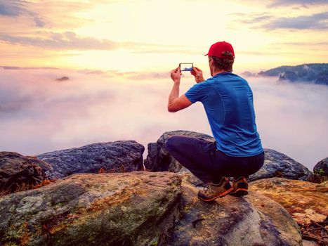 Hiker takes selfie photo, fall nature adventure. Man sit on stone on mountain sumit  Daybreak horizon above milky fog.