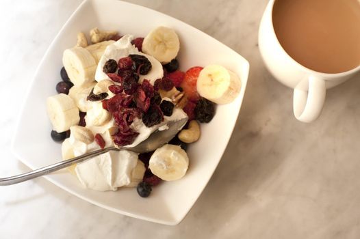 Healthy breakfast serving of assorted fruit with blueberries, strawberries , sliced fresh banana and raisins on fresh creamy yoghurt with tea