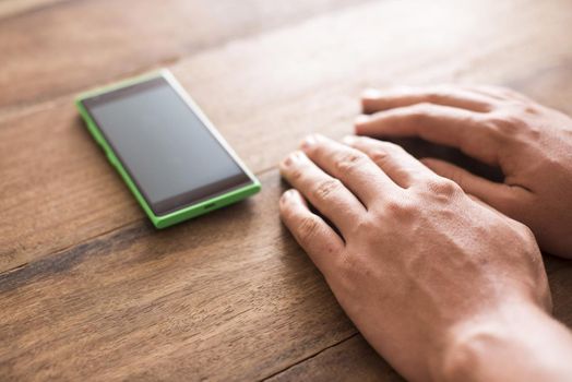 Hands of a man resting on a wooden table alongside his mobile phone as he waits impatiently for a long-awaited call