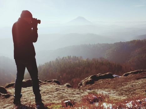 Professional photographer takes photos with mirror camera on peak of rock. Dreamy fogy landscape, spring orange pink misty sunrise in beautiful valley below. Professional photographer.