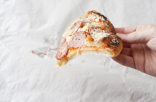 Hand of a man holding a single slice of traditional pepperoni salami Italian pizza over a crinkled sheet of white paper with copy space
