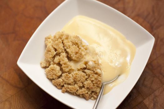 Portion of apple crumble with custard in a modern square bowl viewed from above on a wooden table