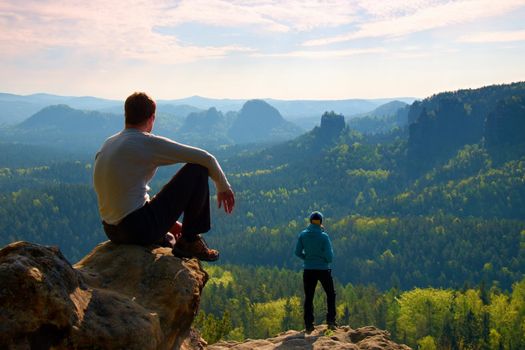 Boy and girl  tourists  stay on cliff and thinking. Dreamy fogy landscape, blue misty sunrise in a beautiful valley below