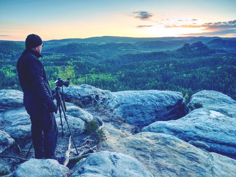 Photographer on a mountain cliff taking picture of landscape awaking.   Dreamy fogy landscape spring orange pink misty sunrise in a beautiful valley below.