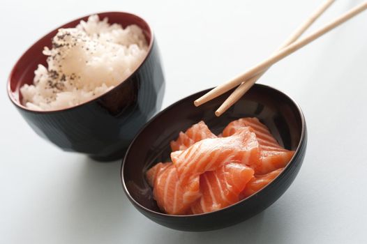 One black bowl of cubed pink salmon and another of steamed rice beside chopsticks against a white background
